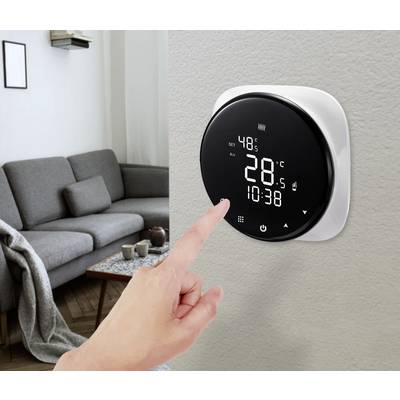 Un doigt touche un thermostat numérique sur le mur d'un salon. L'écran affiche 28°C et l'heure 10:38.