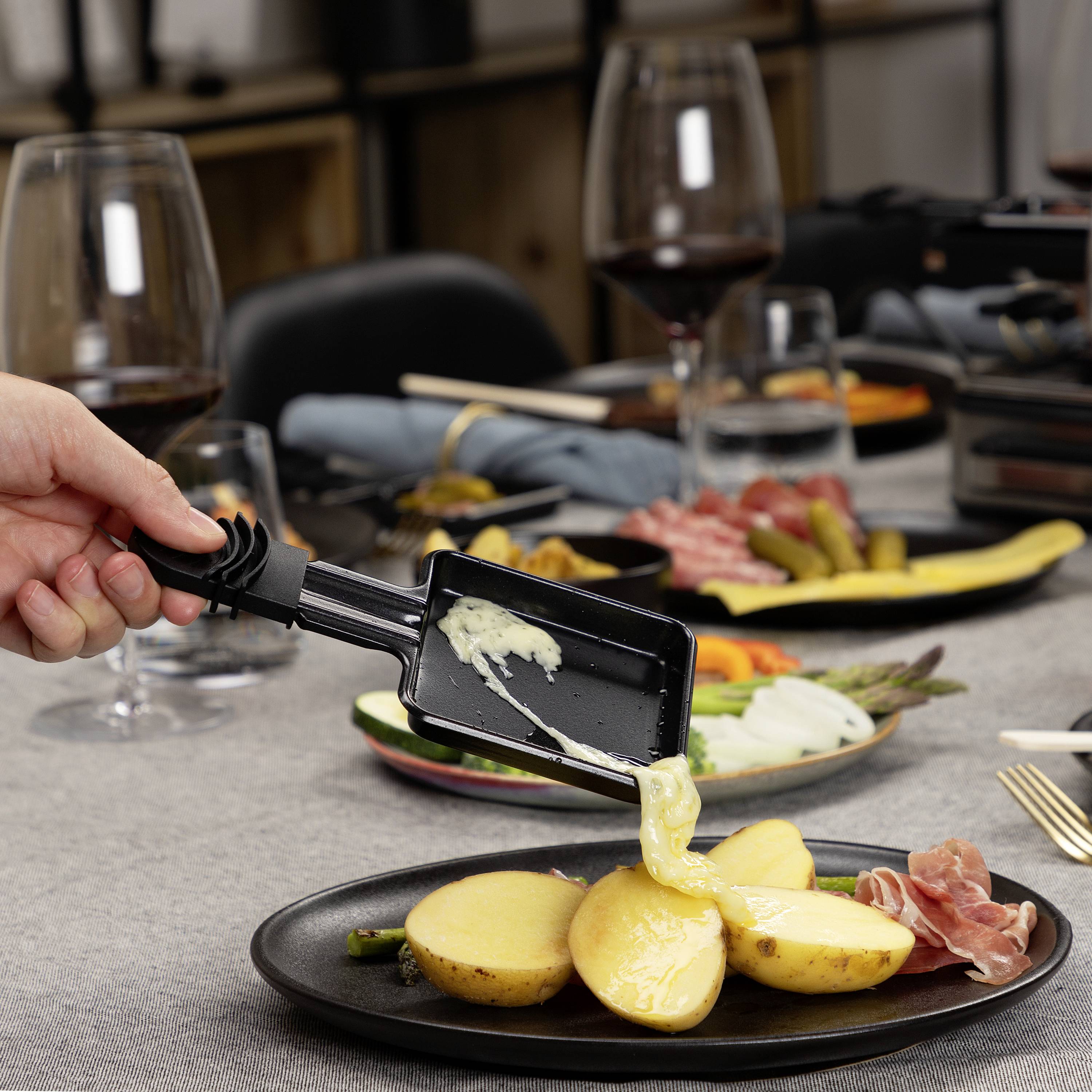 Une main verse du fromage fondu d'un petit plat à raclette sur des pommes de terre dans une assiette. À l'arrière-plan, du vin et des accompagnements.