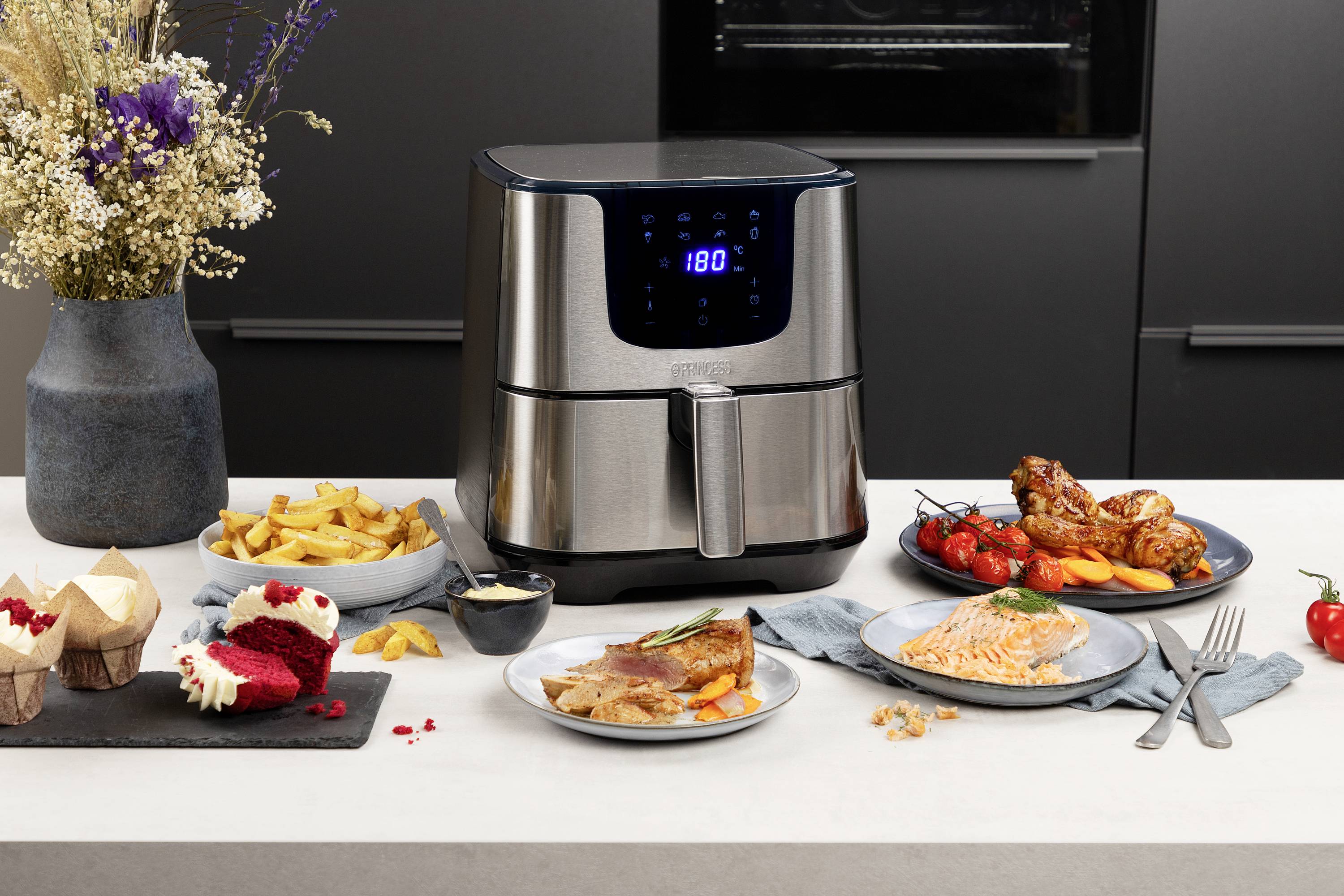 Un air fryer est posé sur un plan de travail de cuisine, entouré de frites, de gâteau, de cuisses de poulet et de plats de poisson.