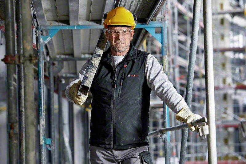 Un ouvrier portant un casque jaune et des gants transporte une pièce métallique sur un chantier de construction. Entouré d'échafaudages et d'équipements de construction.