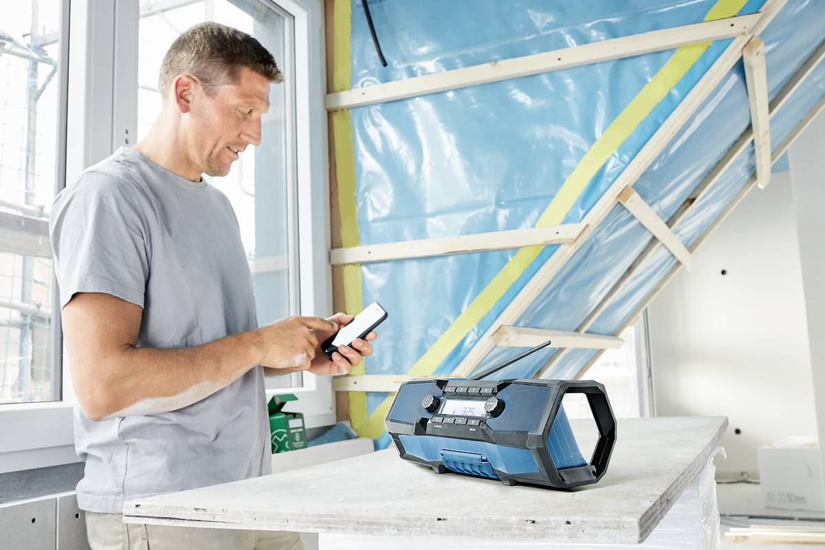 Un homme dans un environnement de construction utilise un smartphone. Un radio portable est posé sur la table devant lui. L'arrière-plan montre un mur de chantier.