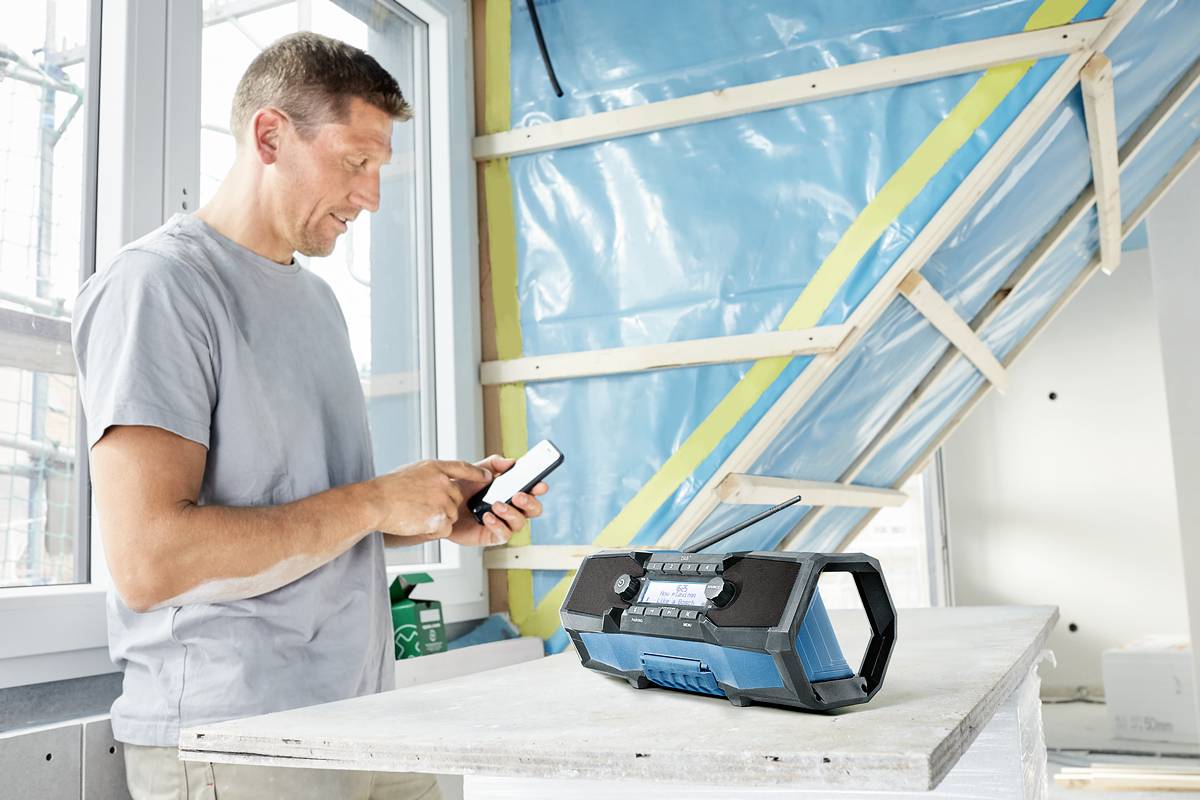Un homme se tient dans une pièce en travaux, utilisant son smartphone. Sur la table devant lui se trouve un radio portable.