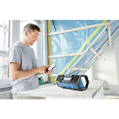 Un homme se tient dans une pièce en travaux, utilisant son smartphone. Sur la table devant lui se trouve un radio portable.