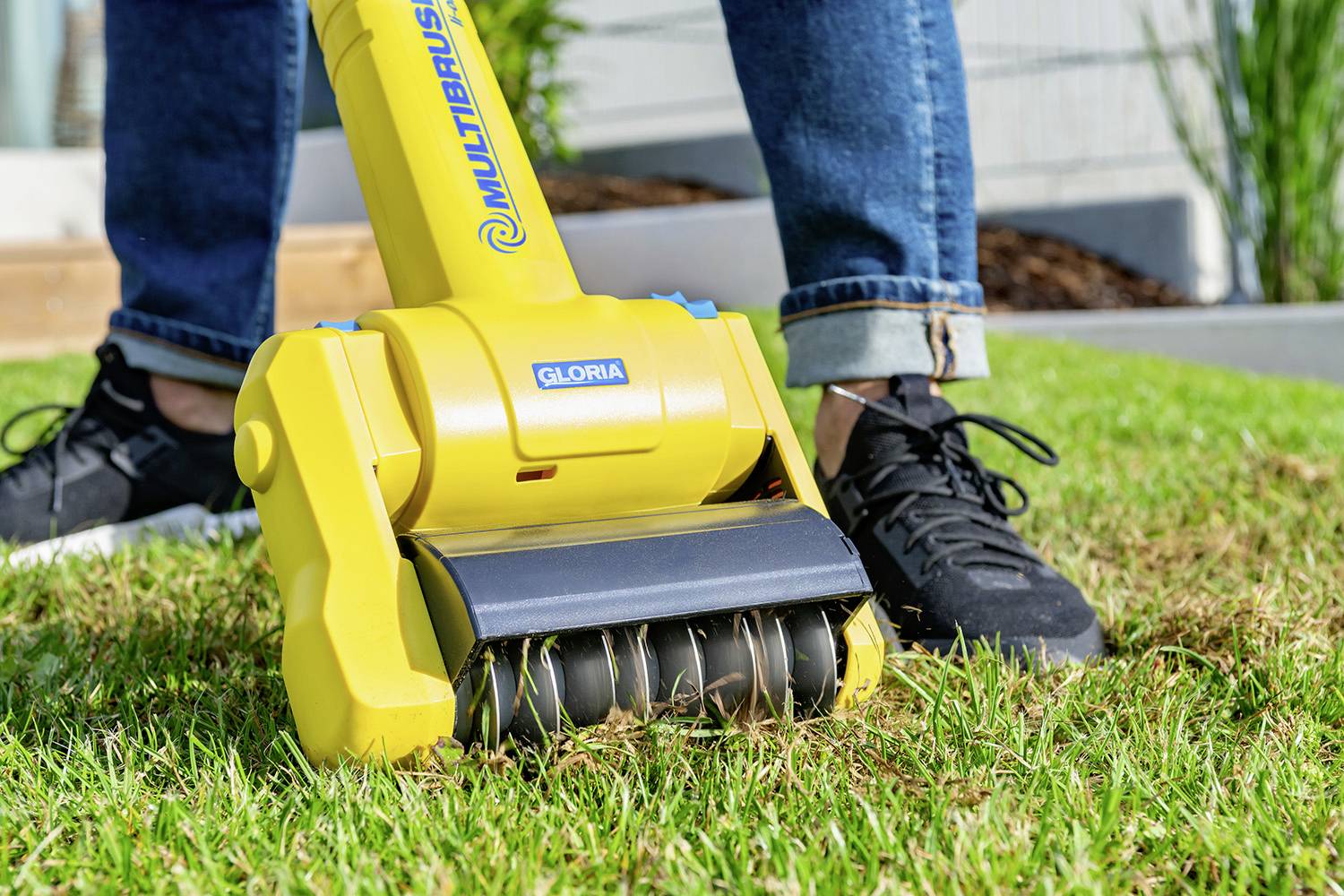 Une personne utilise un aérateur électrique pour gazon sur une pelouse verte. L'appareil a un boîtier externe jaune et des rouleaux noirs.
