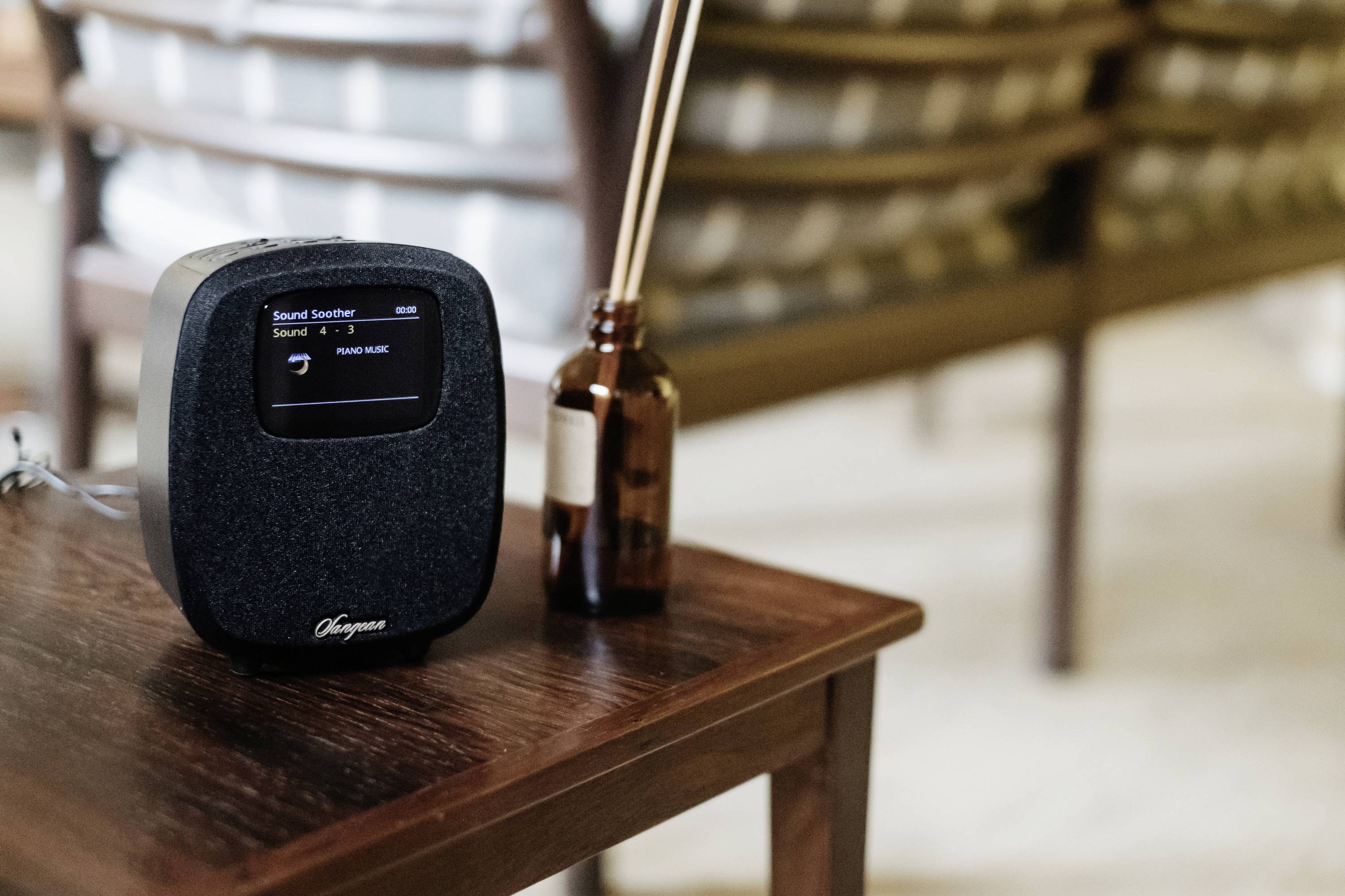 Un haut-parleur posé sur une table en bois affiche « Stand by » sur son écran. En arrière-plan, flou, des chaises et une petite bouteille brune.