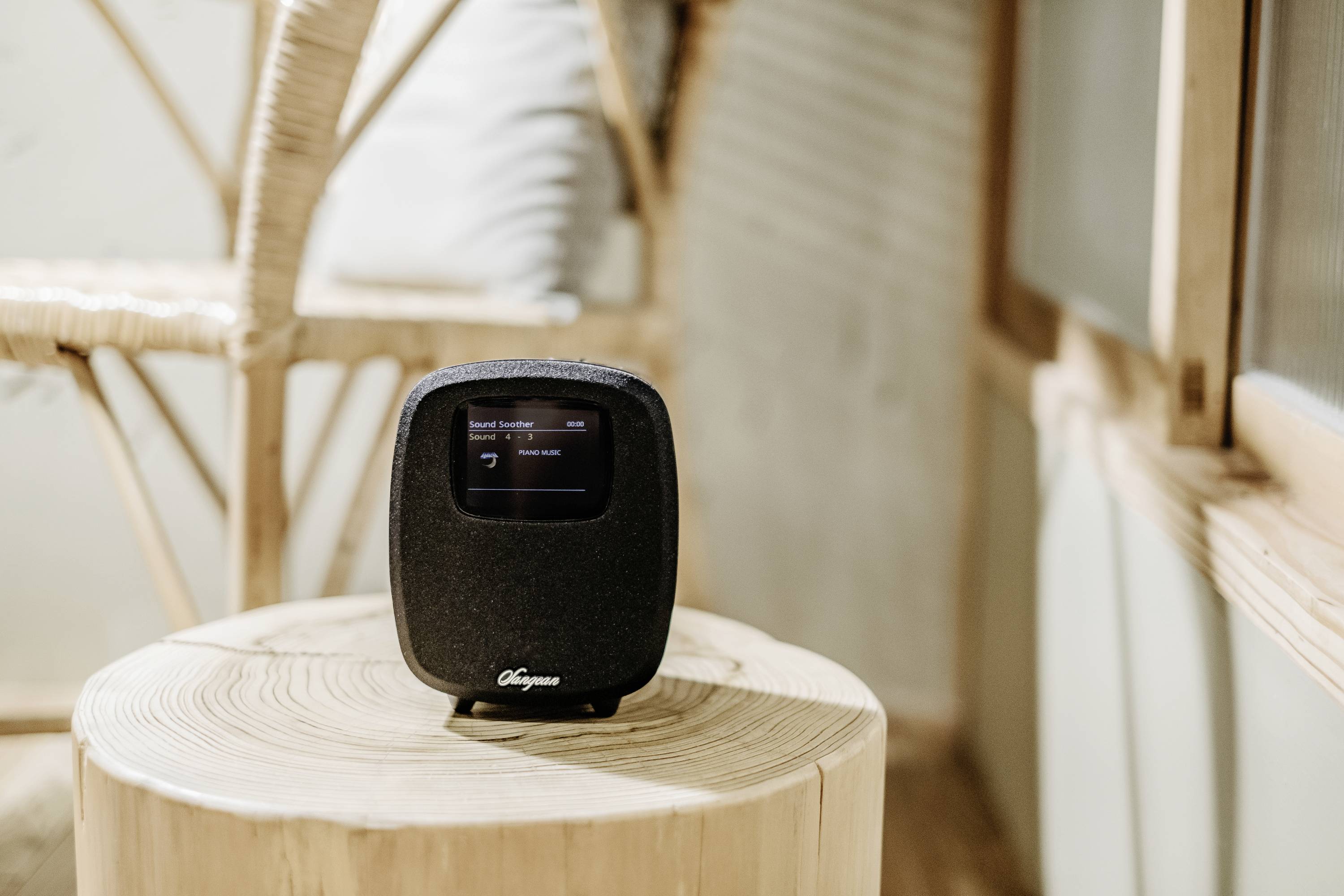 Petit haut-parleur noir avec écran posé sur une table en bois, affichant un titre musical. Un fauteuil en rotin est visible en arrière-plan.