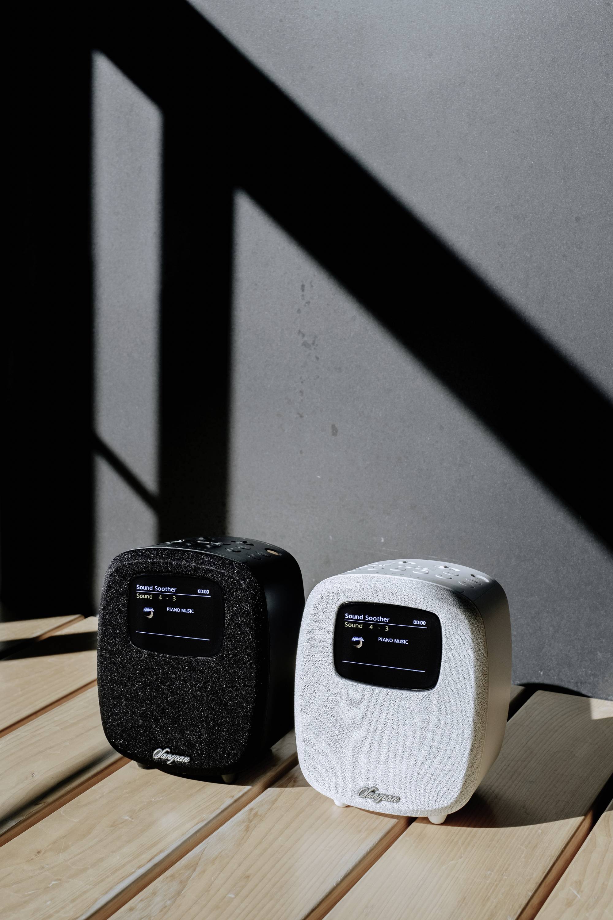 Deux enceintes portables, l'une noire et l'autre blanche, sont placées côte à côte sur une table en bois, baignées de lumière et d'ombres claires.