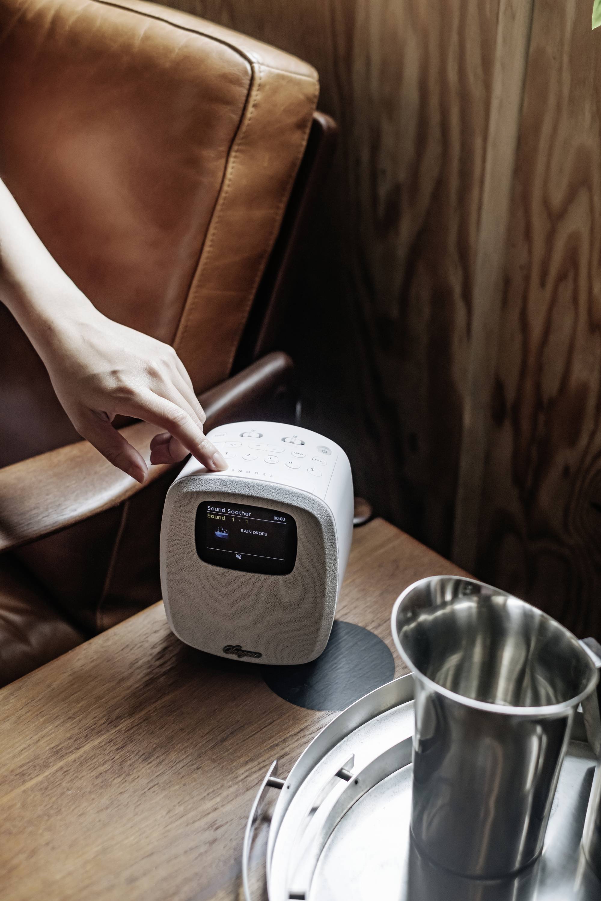 Une personne tape sur un appareil audio compact et moderne posé sur une table en bois, à côté d'un fauteuil en cuir et d'un pichet en métal.