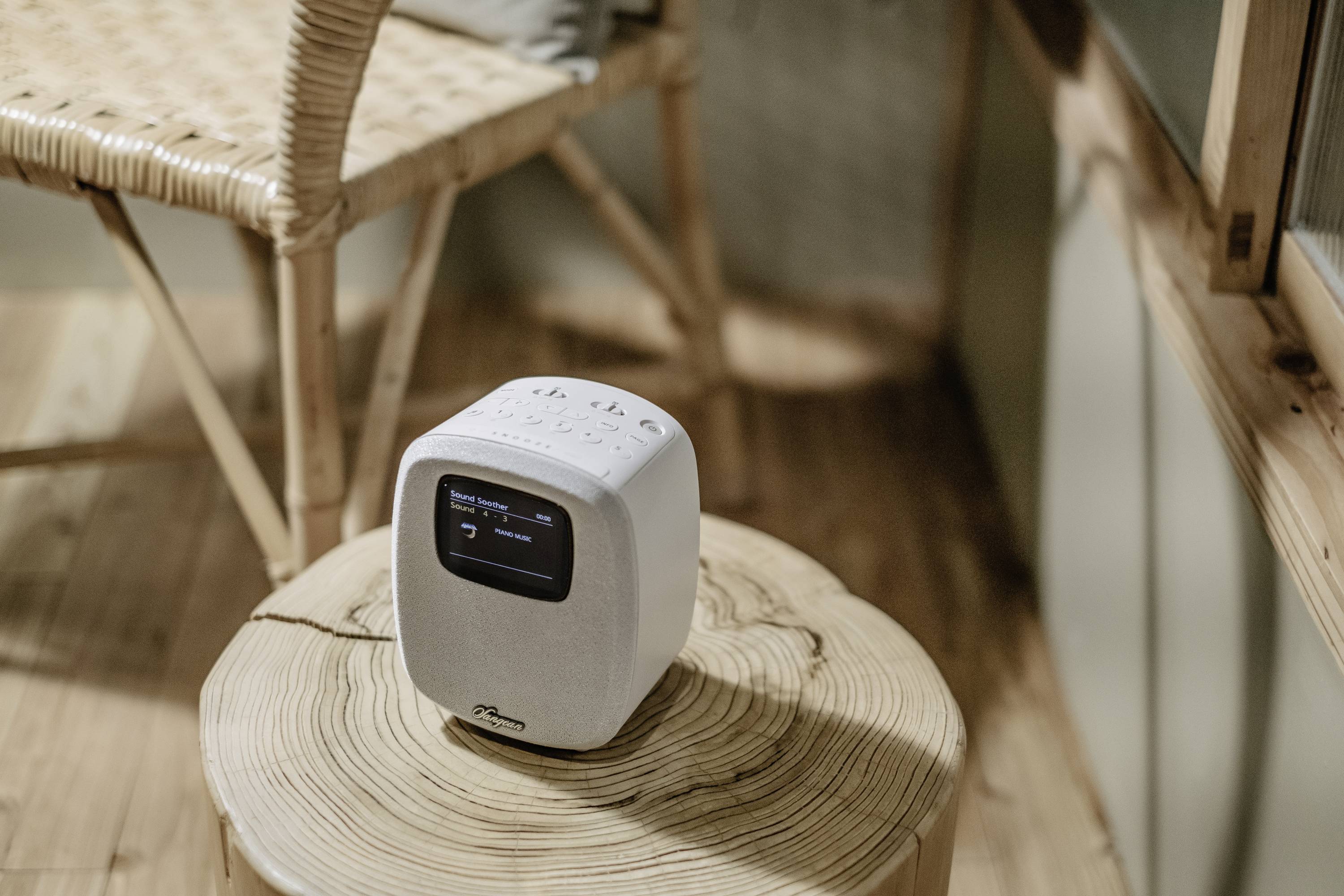 Un petit moniteur de qualité de l'air blanc est posé sur une table ronde en bois dans une pièce au sol en bois avec une chaise en osier. Un affichage numérique montre des données.