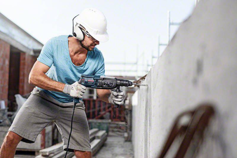Un ouvrier portant un casque de sécurité et des protections auditives fore un mur en béton avec une perceuse sur un chantier de construction.