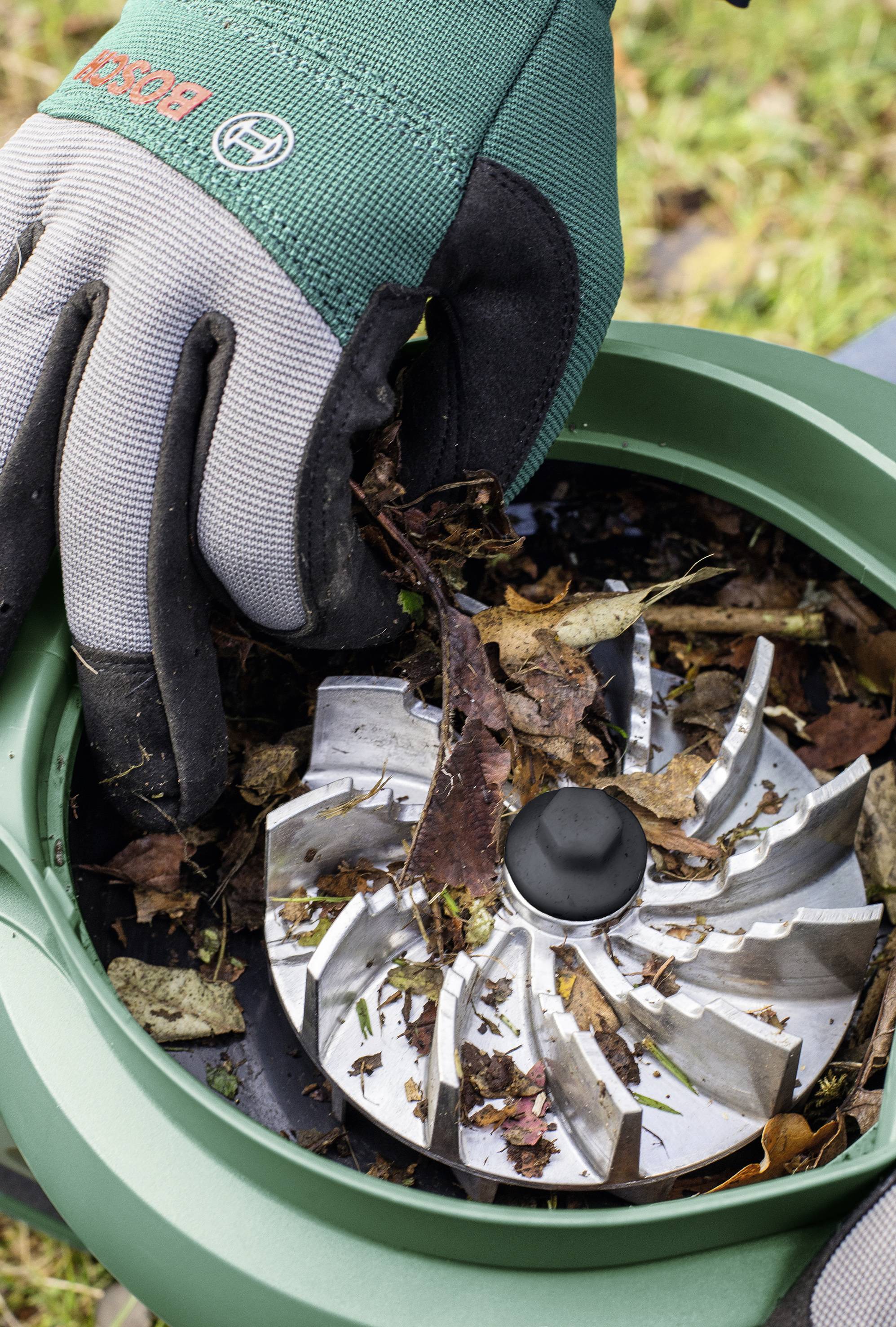 Une main gantée place du matériel végétal dans un broyeur de jardin équipé de lames métalliques. Traitement des déchets verts.