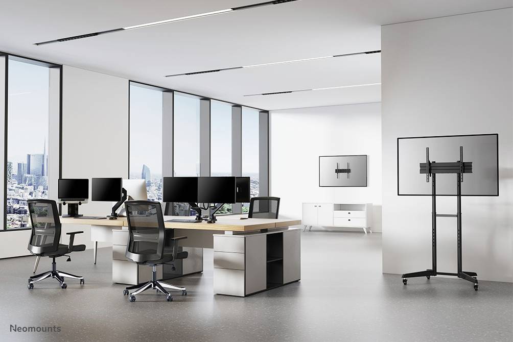 Bureau moderne et lumineux avec de grandes fenêtres. Table centrale équipée de plusieurs écrans d'ordinateur et de chaises pivotantes. Grand écran sur le côté.