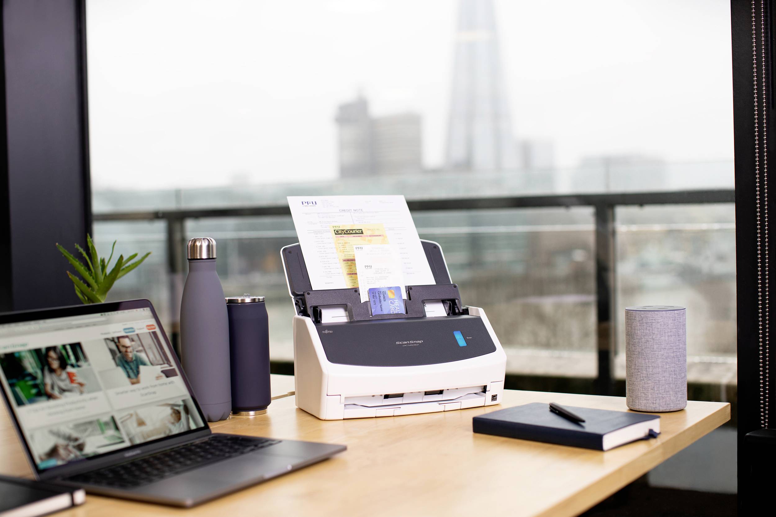 Un scanner sur un bureau numérise un document. À côté se trouvent un ordinateur portable, une bouteille d'eau, un carnet de notes et un haut-parleur. En arrière-plan, on aperçoit la silhouette floue d'un gratte-ciel.