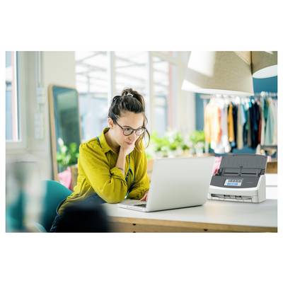 Une femme dans un bureau moderne et bien éclairé utilise un ordinateur portable sur un bureau blanc. Une imprimante est placée à côté d'elle. Des portants à vêtements sont visibles en arrière-plan.