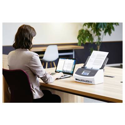 Une femme utilise un ordinateur portable sur un bureau, avec un scanner à alimentation automatique à sa droite, en train de numériser des documents. Environnement de bureau avec un autre bureau à proximité.