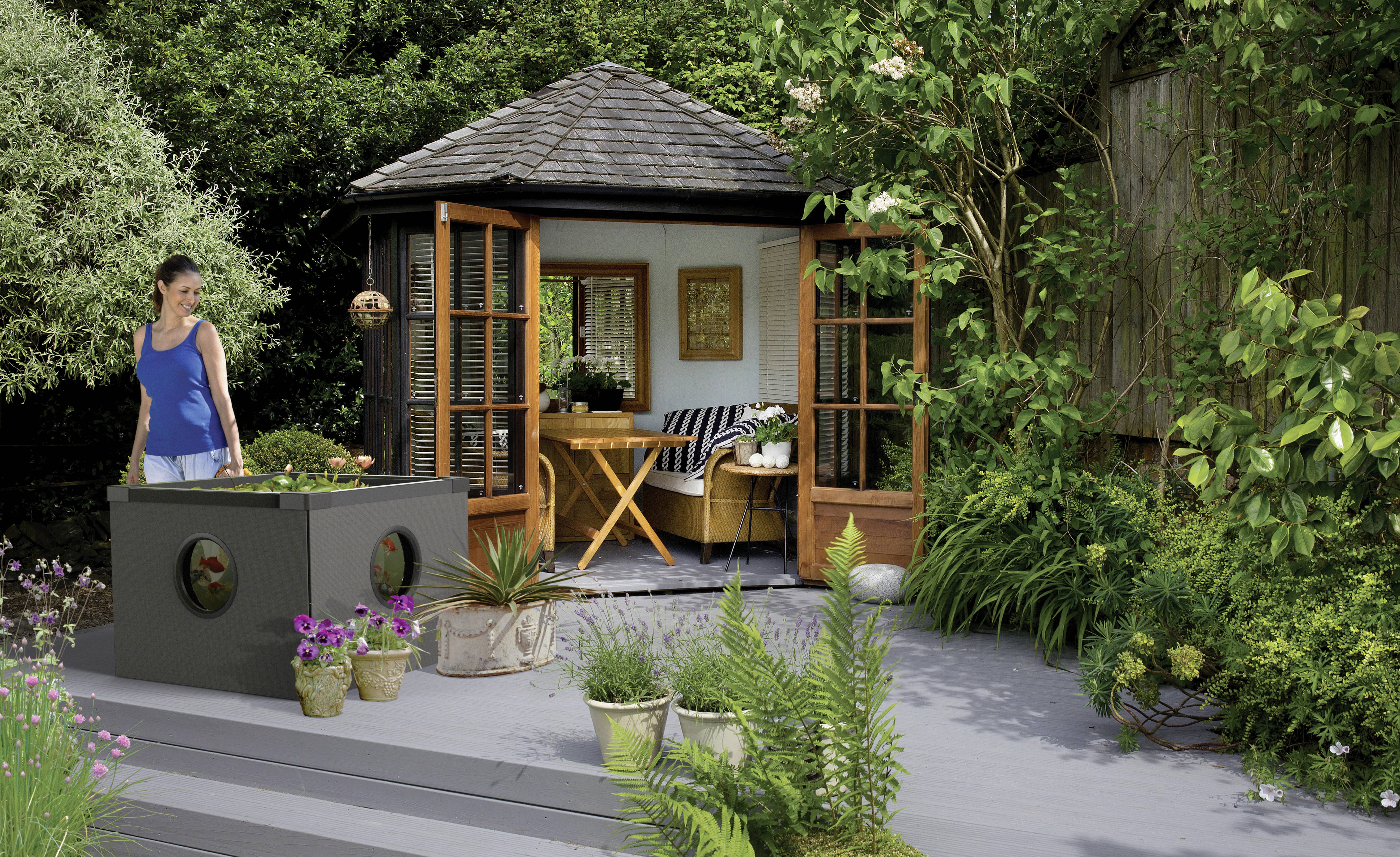 Femme en robe bleue debout dans un jardin ensoleillé avec de nombreuses plantes et une petite maison en bois en arrière-plan.