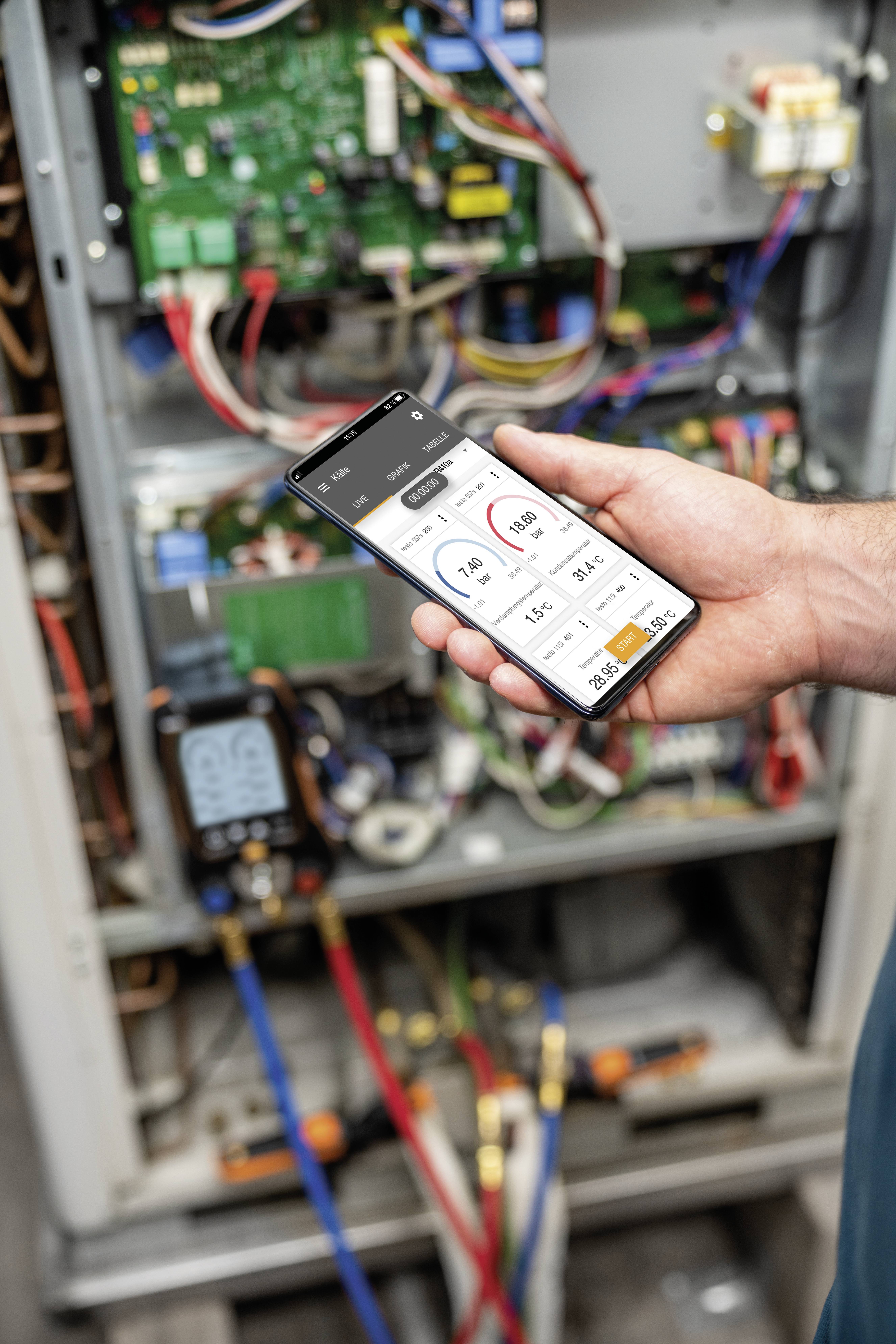 Une personne tient un smartphone avec une application de surveillance de dispositifs devant une armoire électrique ouverte.