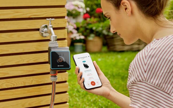 Une femme dans un jardin utilise une application pour smartphone afin de contrôler un système d'irrigation numérique fixé à un mur en bois.