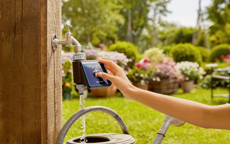 Une personne règle un programmateur d'arrosage sur un robinet, permettant une irrigation contrôlée. L'arrière-plan présente un jardin verdoyant avec des fleurs.