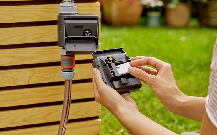 Personne tenant un dispositif de jardinage tout en insérant une batterie, connecté à un tuyau d'arrosage avec un arrière-plan en bois ; scène extérieure.