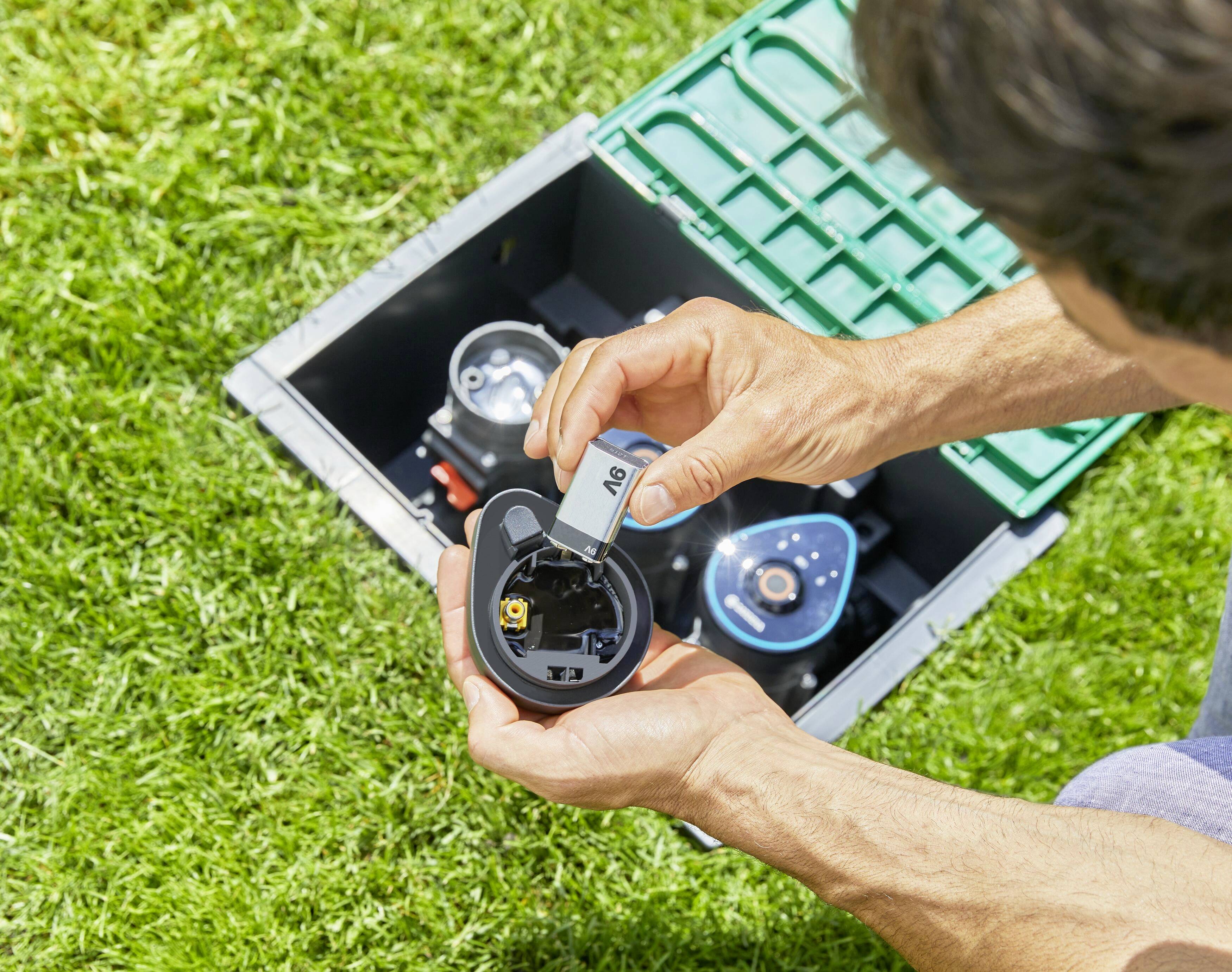 Une personne installe une pile AA dans un appareil électrique à l'extérieur, sur de l'herbe verte, à côté d'un boîtier noir avec des détails verts.