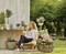 Une femme est assise de manière détendue sur une chaise dans le jardin, entourée de plantes et de fleurs en fleur. À l'arrière-plan, on aperçoit une table avec des pots de fleurs.