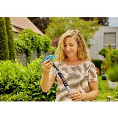 Une femme dans un jardin tenant un petit râteau, entourée de plantes vertes luxuriantes, suggérant des activités de jardinage.
