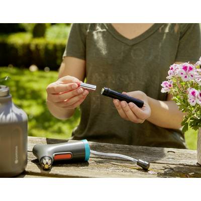 Une personne est assise à l'extérieur et tient un compartiment de batterie d'un appareil portable. Des fleurs et un autre appareil sont visibles sur la table.