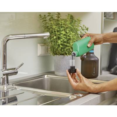 Une personne verse un liquide d'un récipient vert dans un autre récipient au-dessus de l'évier. Une plante est visible en arrière-plan.