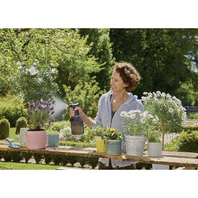 Une femme souriante arrose des fleurs avec un vaporisateur dans un jardin. Différentes plantes en pot sont disposées sur une table en bois à l'extérieur.
