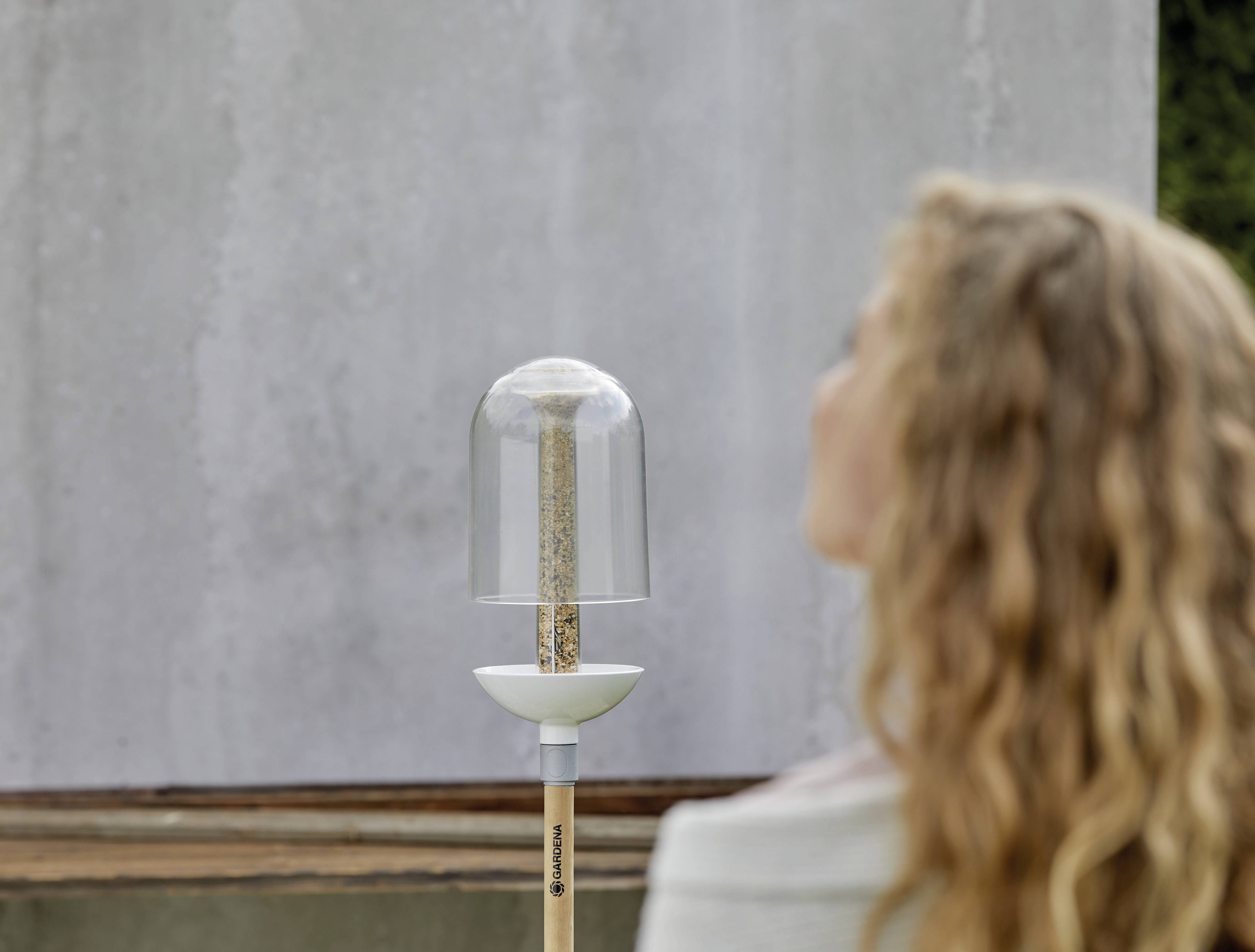 Une personne aux cheveux bouclés observe une mangeoire moderne et transparente fixée sur un piquet, placée devant un mur gris.