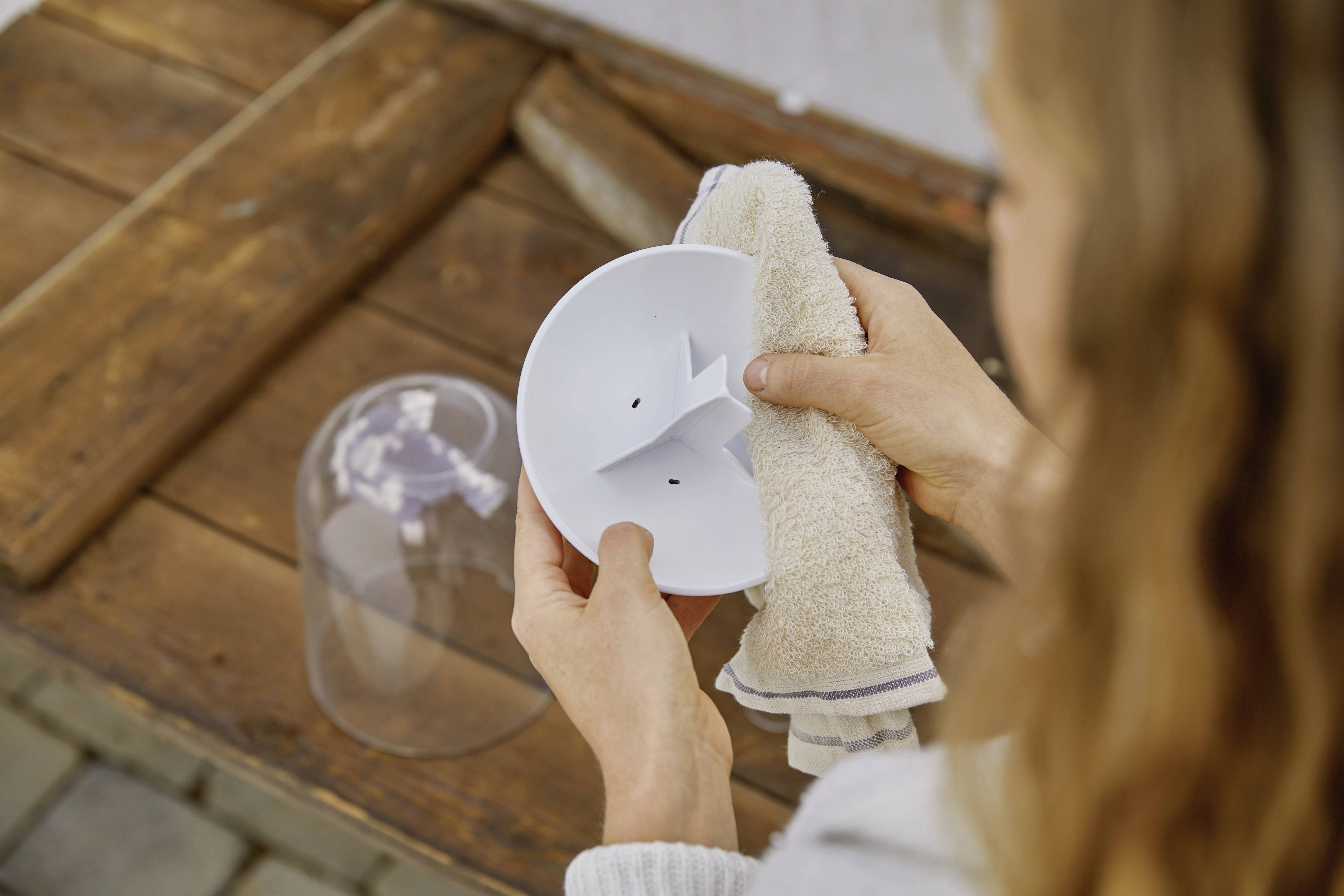 Une personne nettoie un support blanc en plastique avec un chiffon. En arrière-plan, un capot transparent en plastique est posé sur une table en bois.
