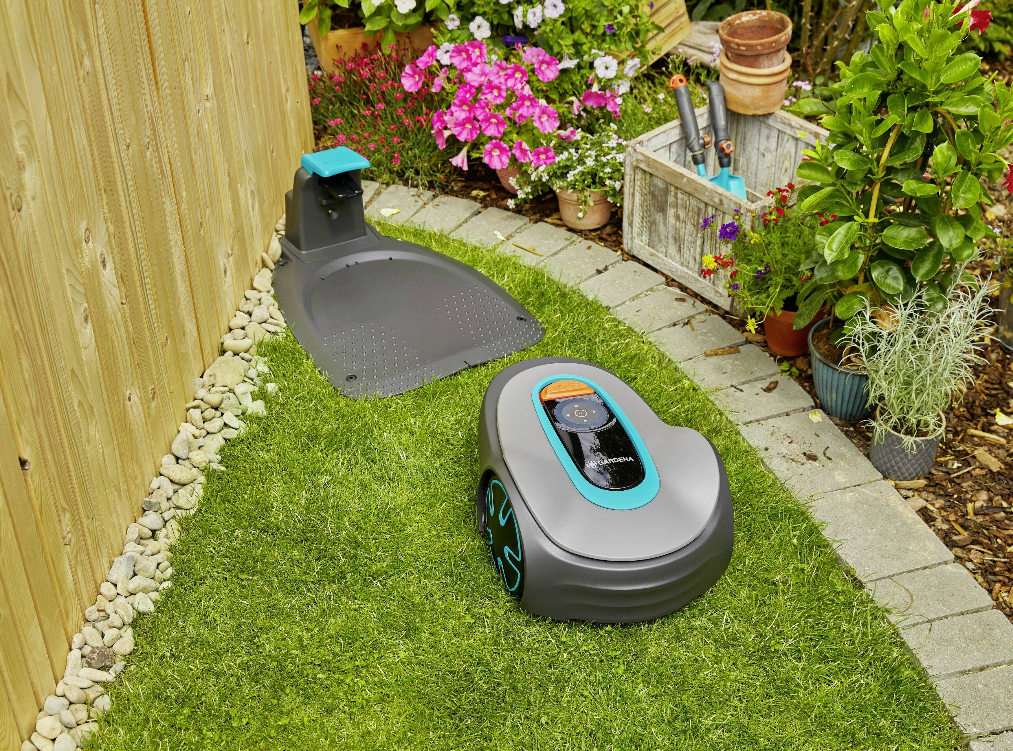 Un robot tondeuse tond la pelouse dans un jardin. À l'arrière-plan, des fleurs colorées sont en fleurs et une étagère en bois avec des outils de jardinage est installée.