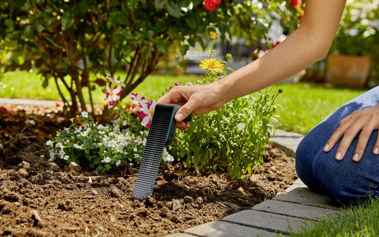 Une personne utilise un testeur de sol dans un jardin, entourée de fleurs en pleine floraison et de verdure, indiquant un intérêt pour le jardinage et la santé du sol.