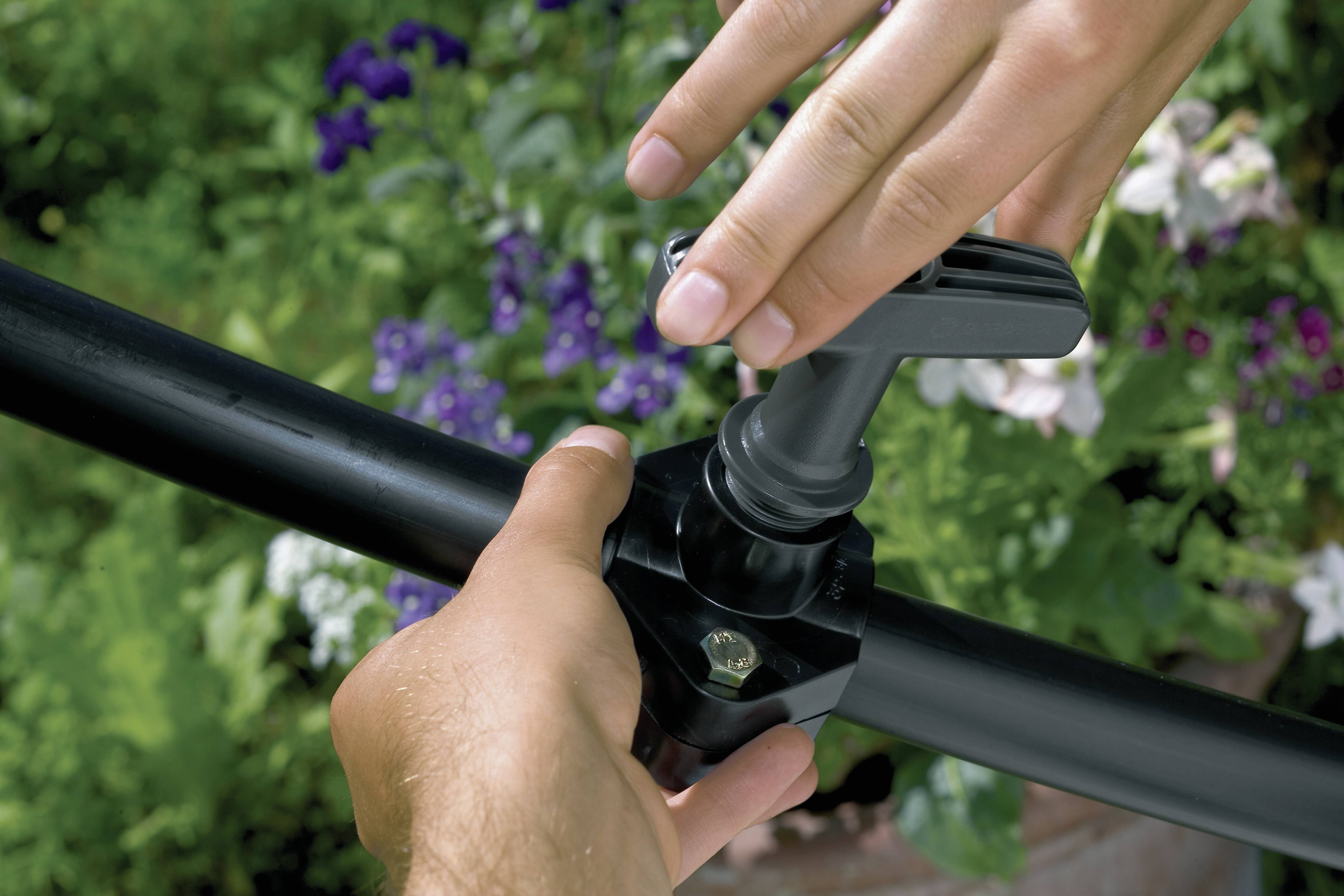 Une personne installe une vanne d'irrigation sur un tuyau noir. En arrière-plan, on aperçoit des plantes vertes floues et des fleurs mauves.