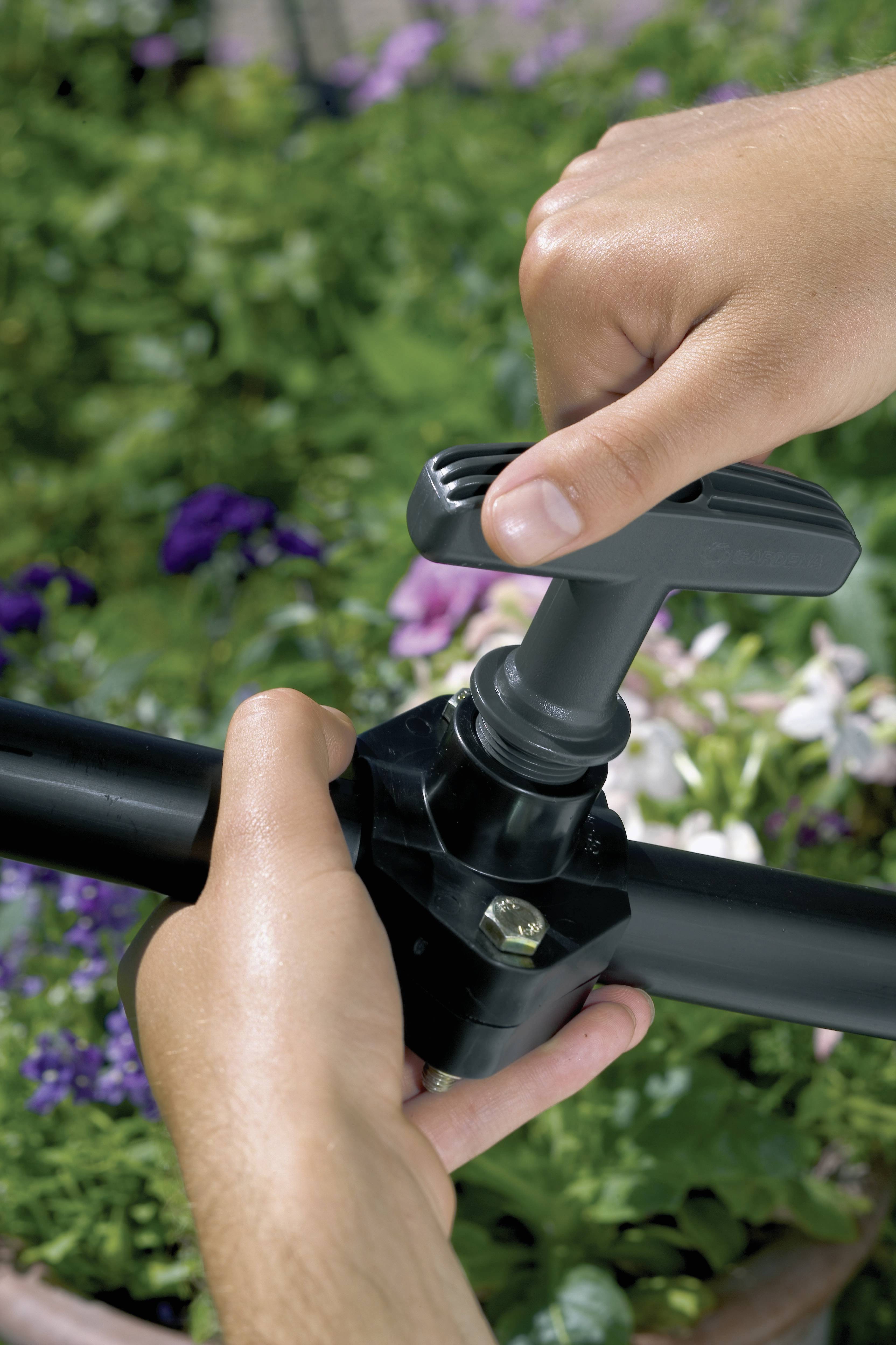Une personne ajuste un robinet de jardin noir à une seule poignée. En arrière-plan, des plantes en fleurs sont visibles.