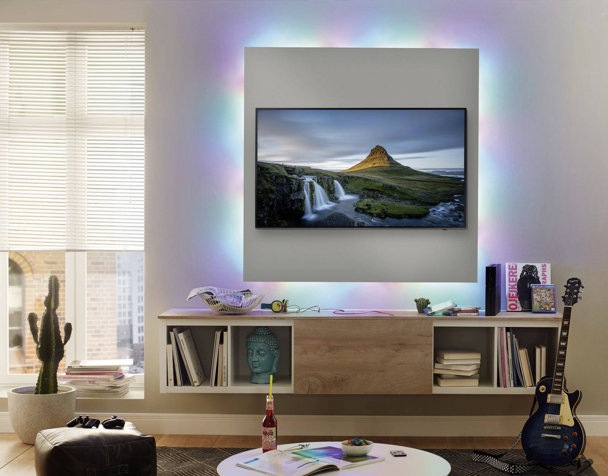 Salon avec un téléviseur mural affichant un paysage. Un éclairage coloré entoure le téléviseur. Une bibliothèque en dessous, avec une guitare à côté.