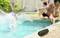 Un groupe de personnes est assis de manière détendue au bord de la piscine. Au premier plan, une personne saute dans l'eau en éclaboussant. Un haut-parleur noir est posé au bord du bassin.