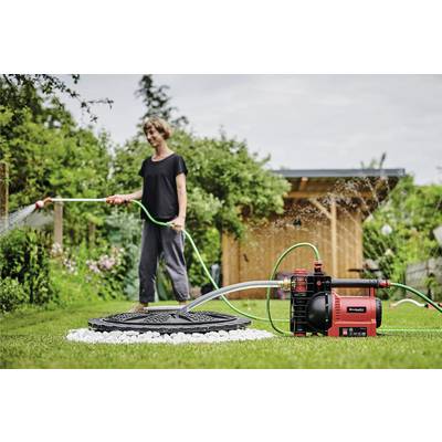 Une personne arrose un jardin à l'aide d'un tuyau connecté à une pompe à eau électrique rouge. Une remise en bois se trouve en arrière-plan.