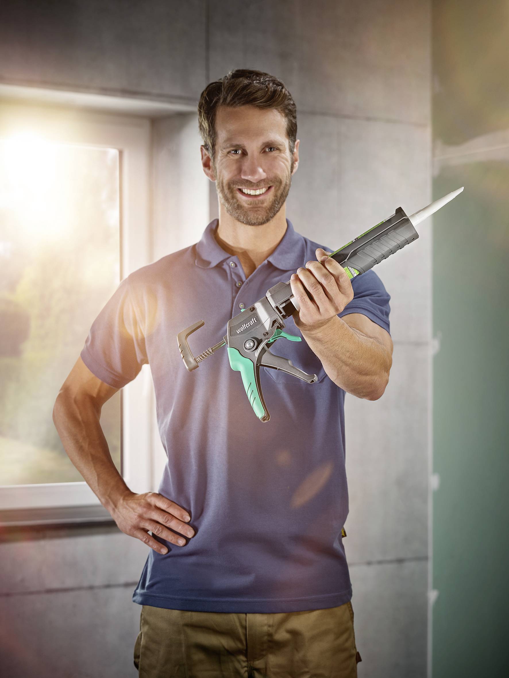 Un homme portant un polo bleu tient un pistolet à mastic dans sa main et sourit dans une pièce lumineuse.