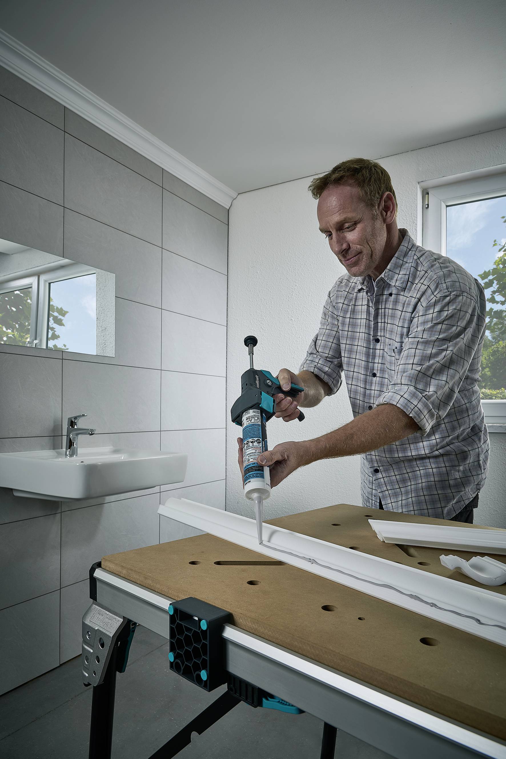 Un homme utilise un pistolet à cartouche pour appliquer de la colle sur un morceau de bois dans une salle de bain. Un lavabo est visible en arrière-plan.