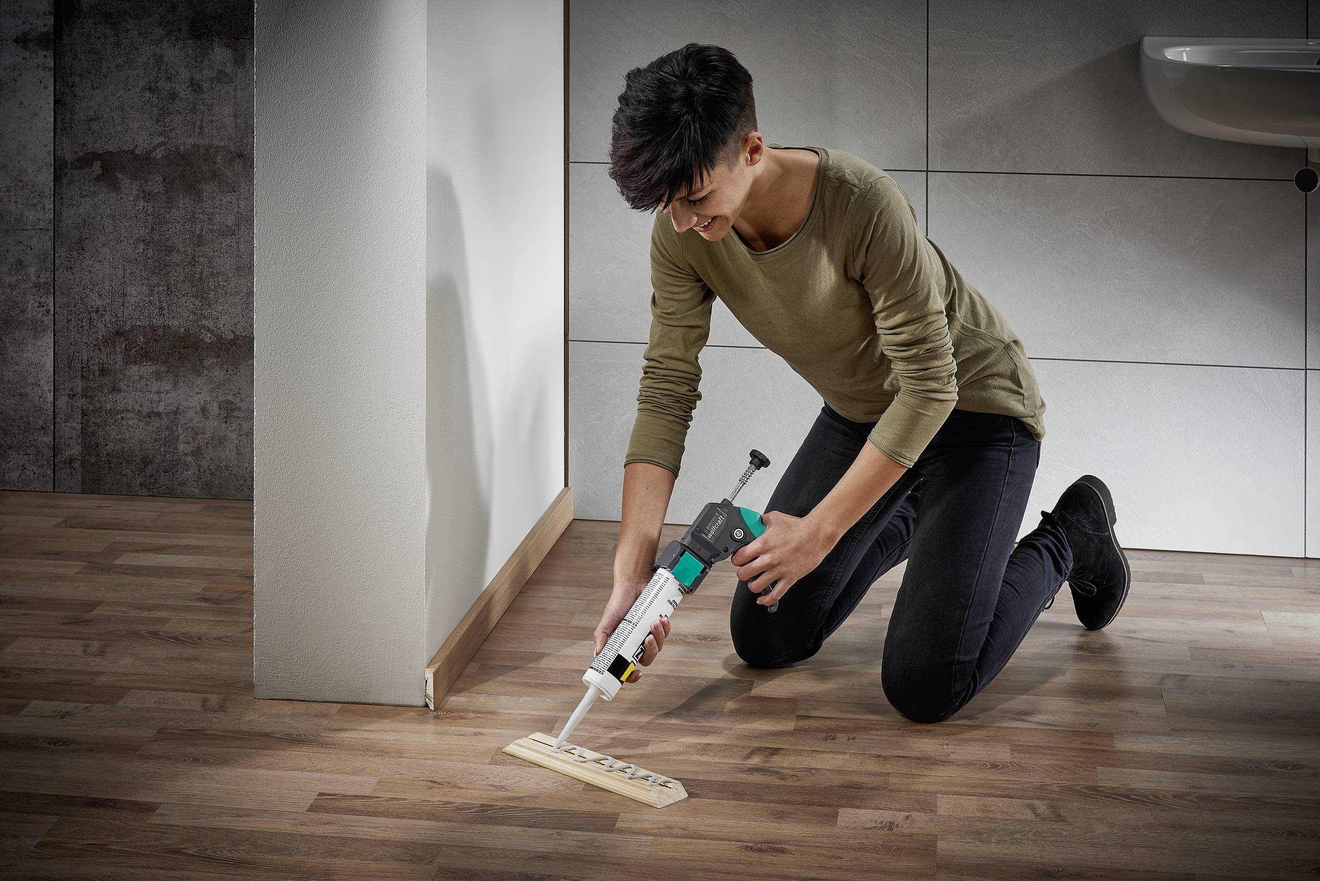 Une personne est agenouillée sur un plancher en bois et utilise un pistolet à silicone pour combler l'espace entre le sol et le mur.
