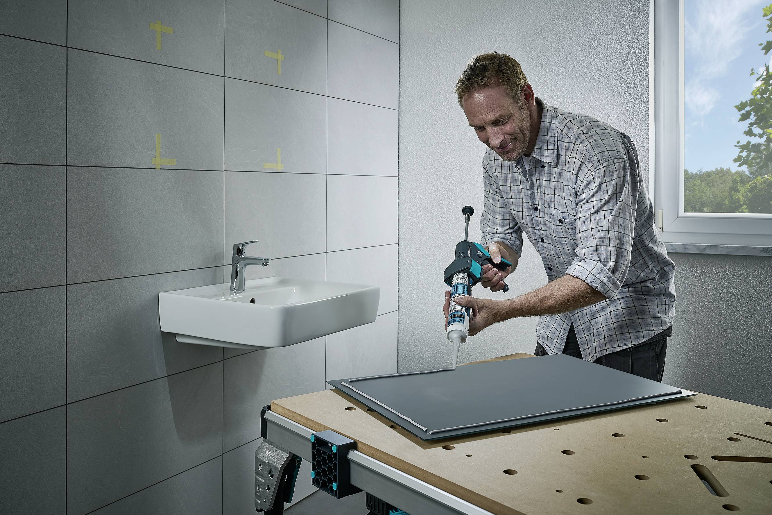 Un homme dans une pièce lumineuse applique un mastic sur une plaque posée sur une table de travail. Un lavabo est visible en arrière-plan.