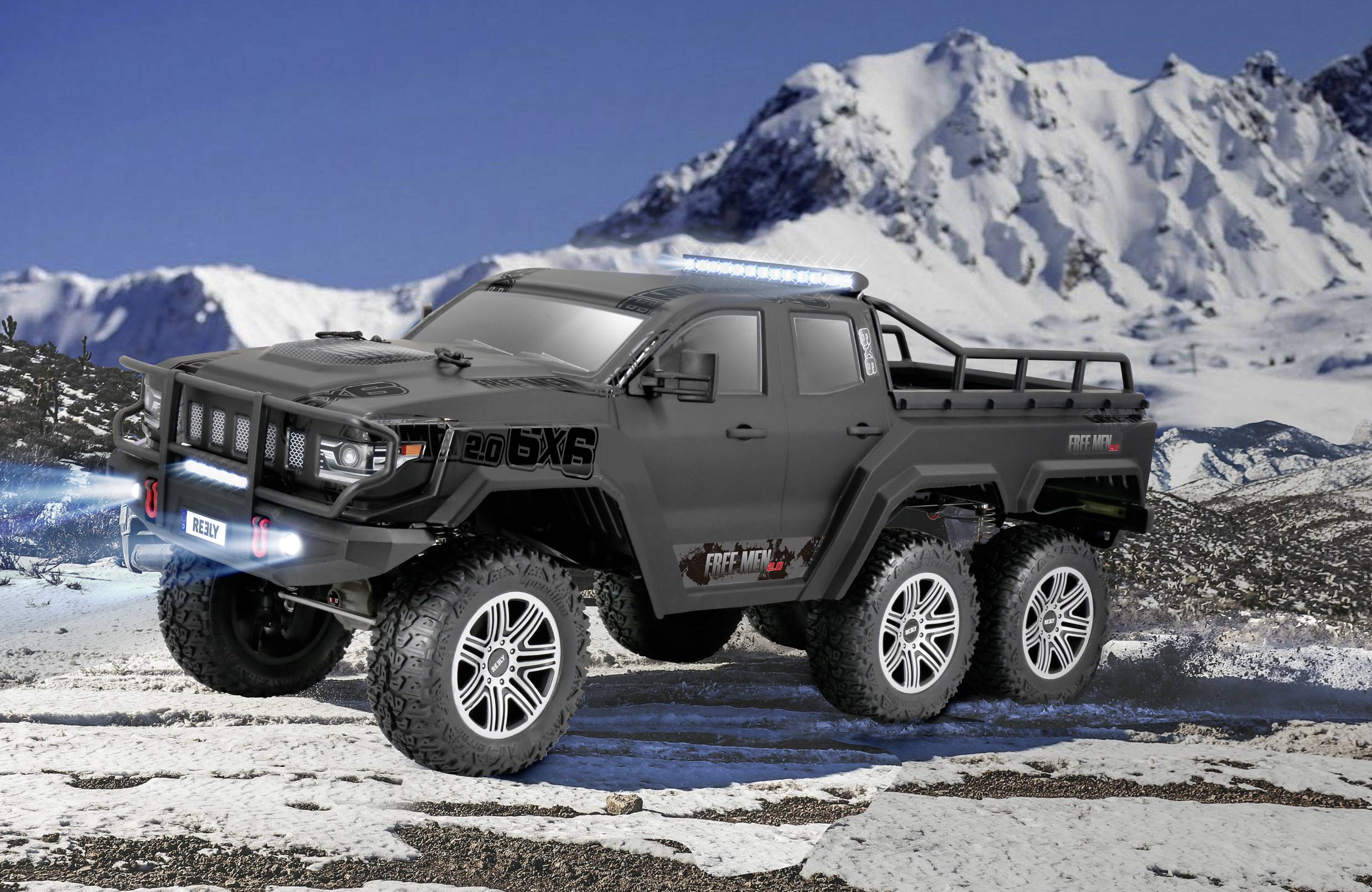 Une voiture miniature noire à six roues est posée sur un terrain enneigé, avec un paysage de montagnes et un ciel bleu en arrière-plan.