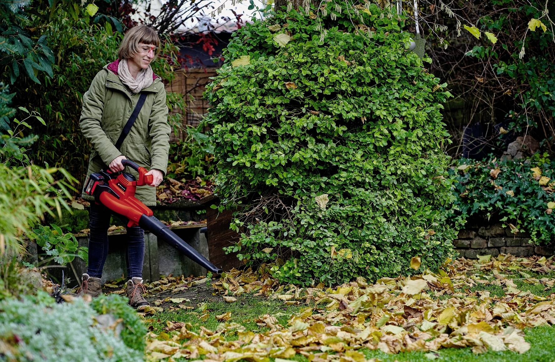 Une personne dans un jardin utilise un souffleur de feuilles rouge pour enlever les feuilles tombées d'une pelouse. Des arbustes et des arbres sont visibles en arrière-plan.