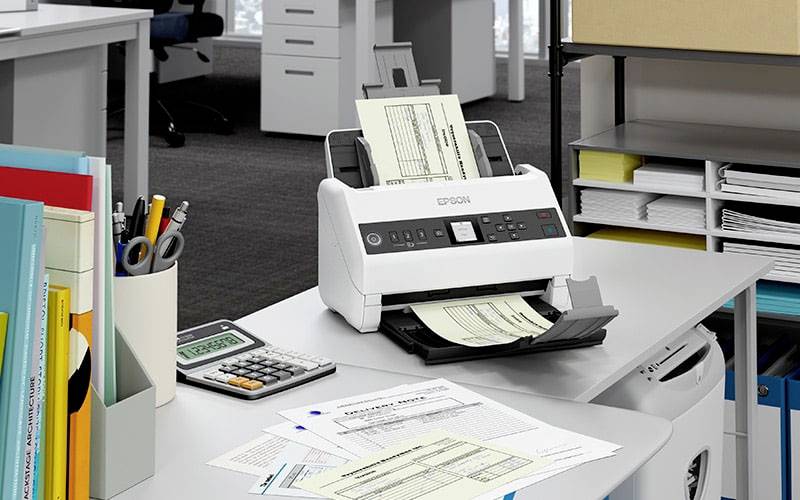 Un scanner de documents sur un bureau numérise plusieurs formulaires. À côté du scanner se trouvent d'autres papiers et une calculatrice.