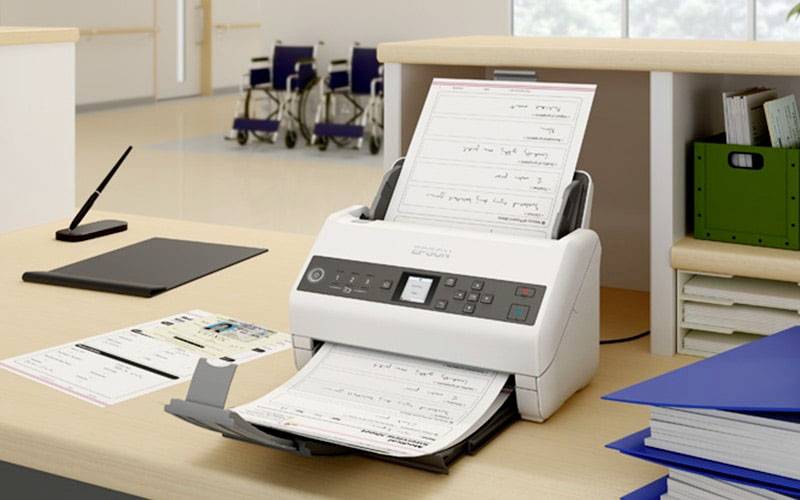 Un scanner numérise des documents dans un bureau. Des classeurs bleus et un porte-bloc noir sont posés sur la table. Des fauteuils roulants sont disposés en arrière-plan.