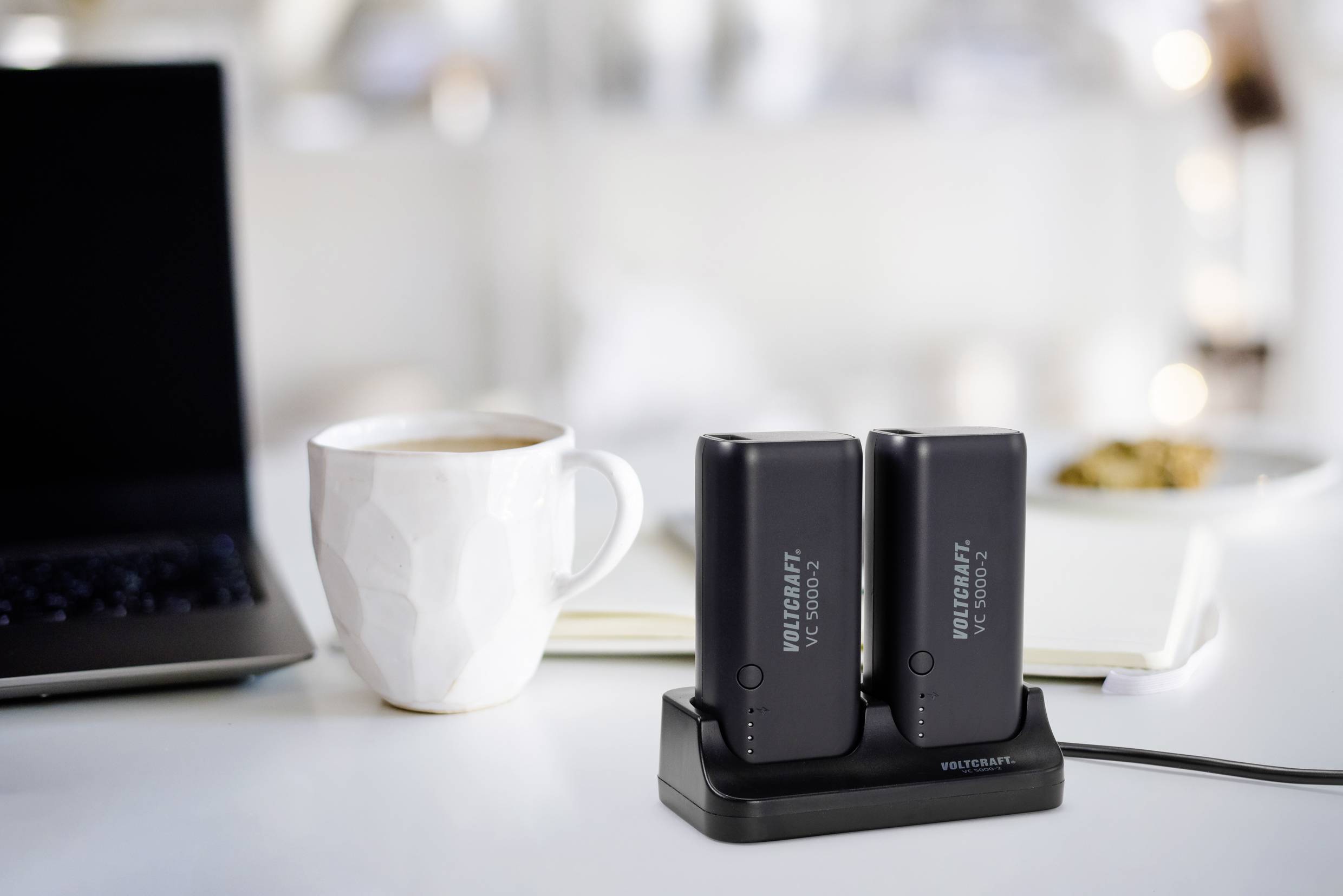 Deux batteries noires sur une station de charge à côté d'une tasse blanche et d'un ordinateur portable ouvert ; ambiance de bureau conviviale.
