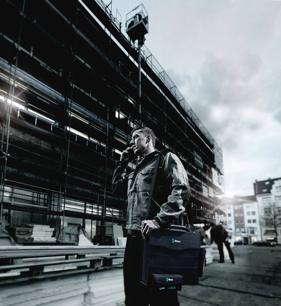 Un homme se tient devant un bâtiment en construction. Il téléphone et porte un sac. Un échafaudage est visible en arrière-plan.