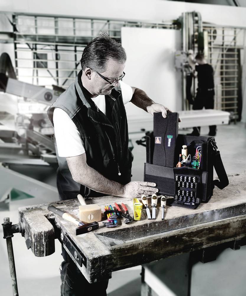 Un artisan dans son atelier ouvre un sac à outils sur un établi, entouré de divers outils et matériaux.